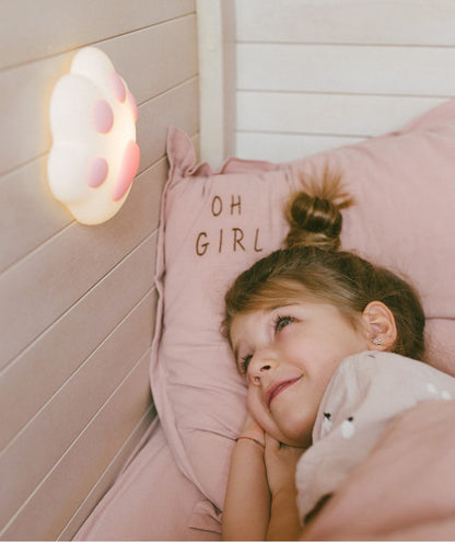 A girl lying in bed with a Paw Silicone Lamp - Pink, featuring a unique design for children's rooms. The lamp offers adjustable light intensity, touch control, and magnet attachment.