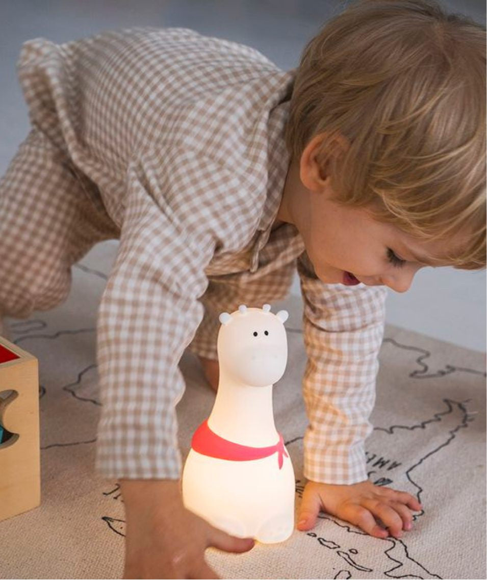 A child plays with a Giraffe Silicone Lamp - White, a friendly night light in the shape of a giraffe with a scarf. Soft, touch-responsive, 7-color lighting, USB rechargeable, safe for kids.