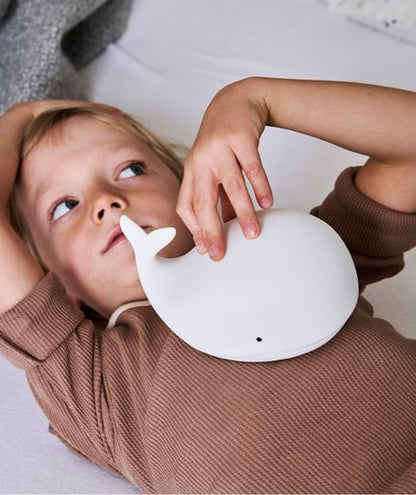 A child lies near a white whale lamp, soft and touchable. The Whale Silicone Lamp with Remote offers 7 colors, touch activation, and safe, BPA-free silicone material for peaceful nights. Dimensions: 8.9 x 13.6 x 10.8 cm.