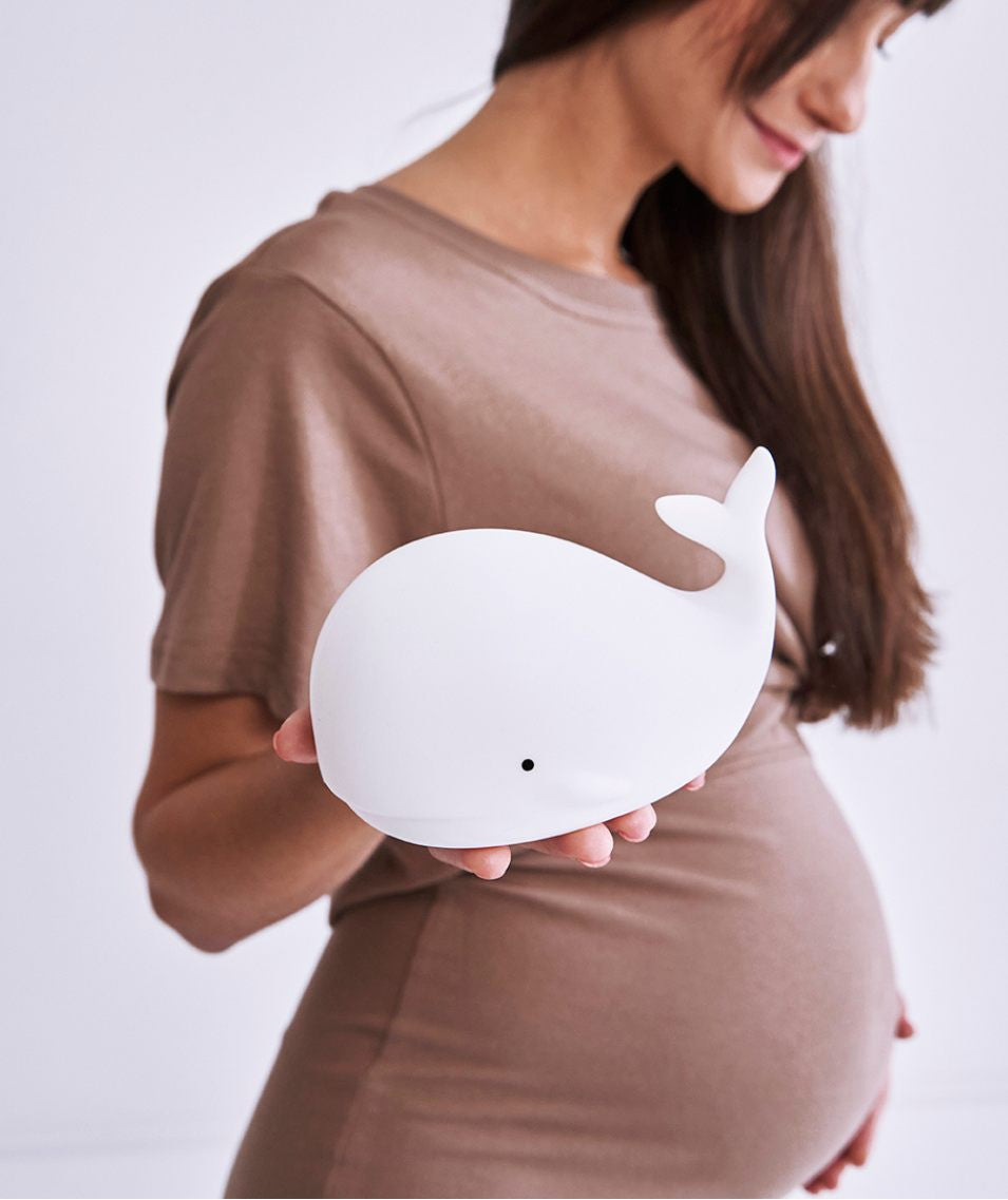 Pregnant woman holding a toy whale, a soft, touchable Whale Silicone Lamp with Remote for kids' safety and comfort. Features 7 colors, patting to light, and 12-hour lighting time. Dimensions: 8.9 x 13.6 x 10.8 cm.