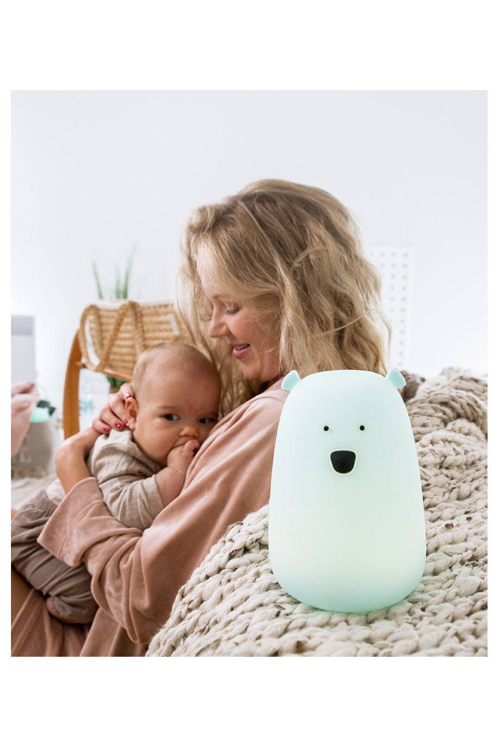 A woman holding a baby, a white object with a face, and a light-up toy on a blanket, embodying the Big Bear Silicone Lamp by Rabbit & Friends, a comforting and playful companion for children's bedtime routines.