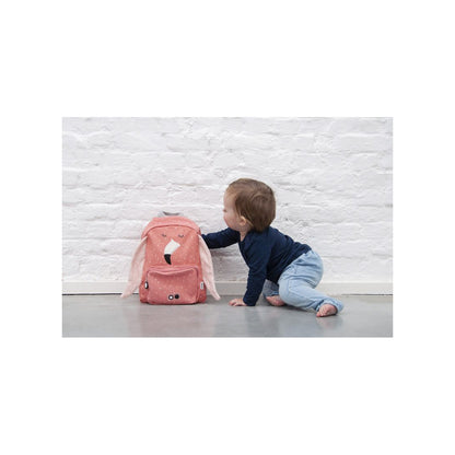 A toddler crawls near a pink Mrs. Flamingo backpack. Adjustable straps, chest strap, water-repellent cotton material, 7.5L capacity, ideal for ages 3+, with front pocket and name tag.