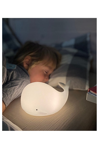 A child peacefully sleeps next to a white whale-shaped lamp, emitting a warm glow. The Whale Silicone Lamp with Remote offers soft touch, color-changing light, and easy pat control for a cozy bedtime experience.